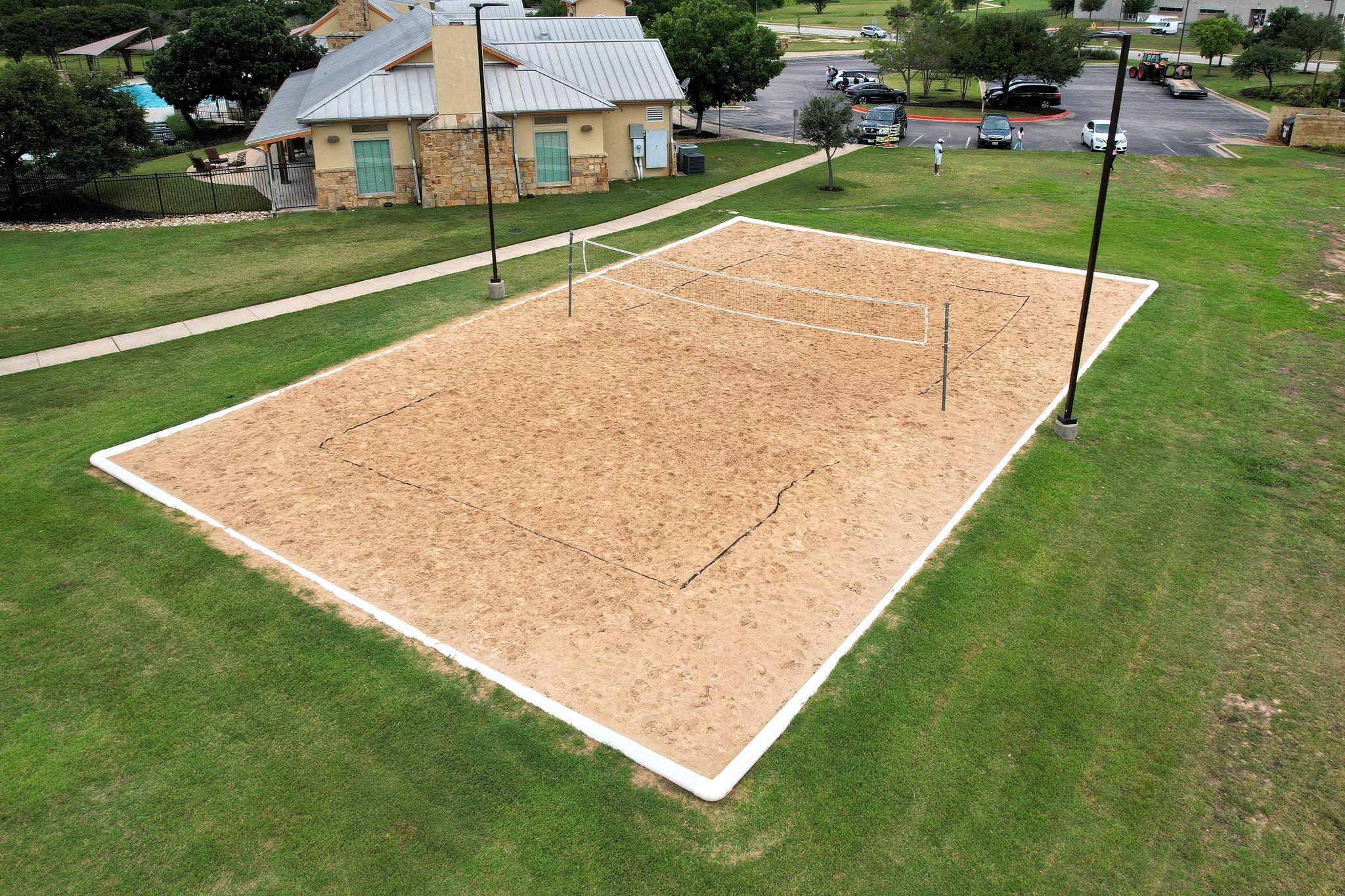 A sand volleyball court surrounded by green grass and light poles, located near the community clubhouse.