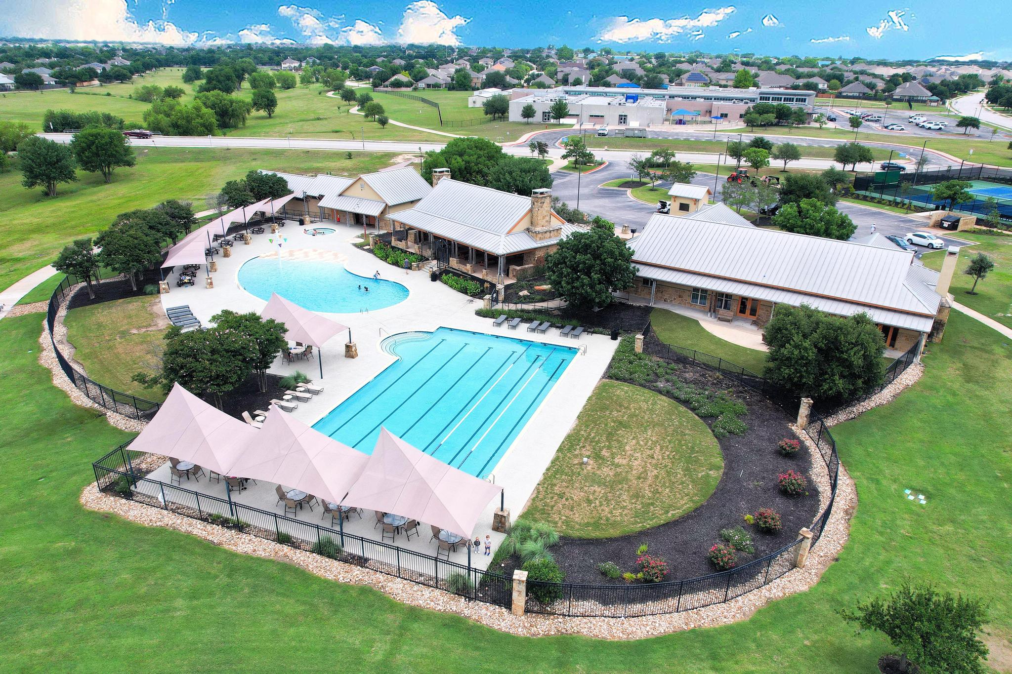 Wide aerial view of a pool complex with lap lanes, shade canopies, and landscaped grounds in a residential neighborhood.