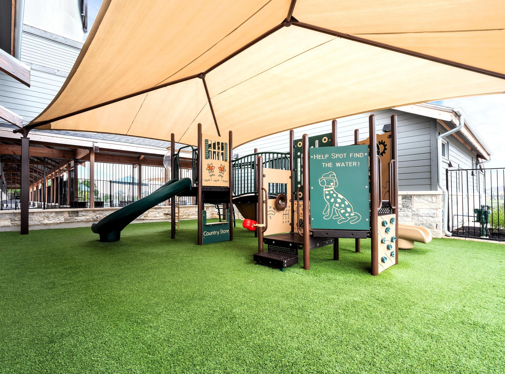 Shaded outdoor playground with artificial turf, slides, climbing panels, and interactive play boards.